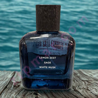 Thumbnail for Perfume bottle labeled 'Lemon Zest, Sage, White Musk' with a wooden cap on a wooden surface and water background