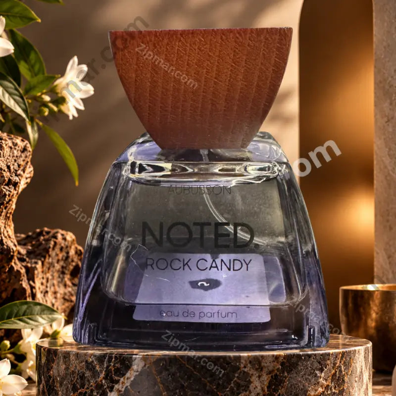 Aubusson Noted. Rock Candy Eau de Parfum bottle with blue-tinted glass and dark wood cap on marble stone pedestal — white jasmine flowers and rough volcanic rocks circular number 2 sticker visible on bottle front warm amber atmospheric lighting
