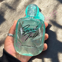 Thumbnail for Hand holding a turquoise 'Sweet Talk' perfume bottle by Jean Marc Paris on a textured surface.