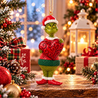 Thumbnail for Enesco Jim Shore Grinch in Santa hat holding heart ornament lifestyle — Grinch in red folk art sweater and green pants clutching large red swirled heart, Christmas background
