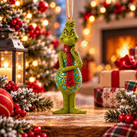 Thumbnail for Enesco Jim Shore Grinch with red scarf ornament lifestyle — standing Grinch with hand on cheek, teal and gold folk art body design, natural jute cord, Christmas scene background