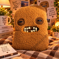 Thumbnail for Fuggler Bigg Nuggets plush with sleepy eyes and gap toothed mouth styled on a plaid blanket with books and a mug”