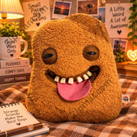 Thumbnail for Fuggler Bigg Nuggets plush with sleepy eyes and pink tongue styled on a plaid blanket with books and a mug”
