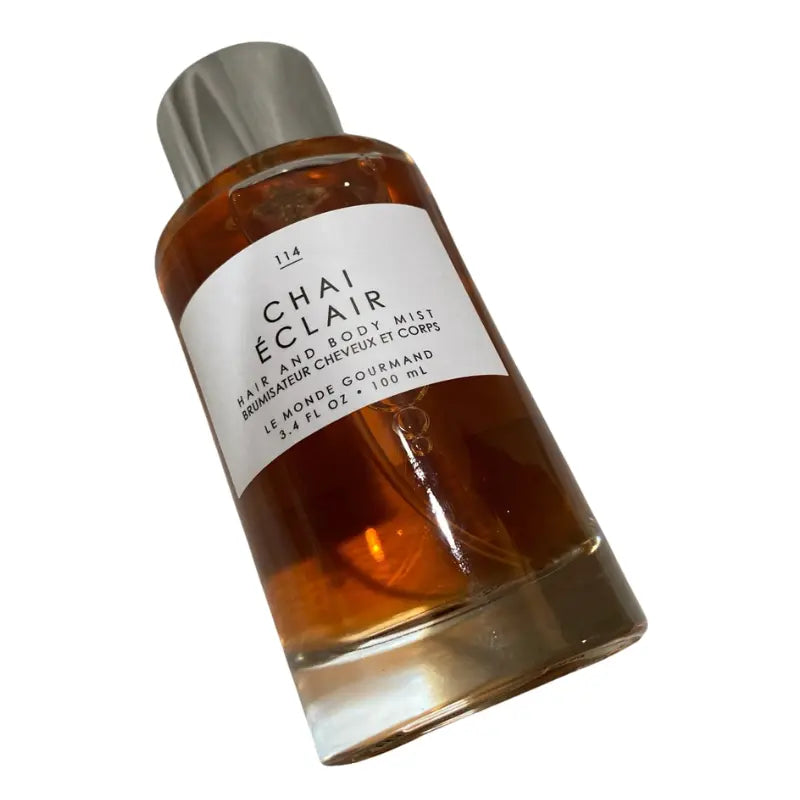 Tilted front view of Le Monde Gourmand Chai Éclair Hair and Body Mist 3.4 oz bottle, highlighting the warm amber-colored liquid.
