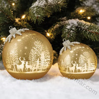 Thumbnail for Two gold glass ornament decor pieces with white reindeer and winter forest scene resting in snow beside a Christmas tree