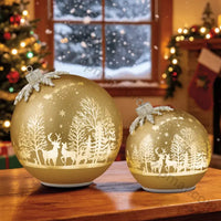 Thumbnail for Set of 2 gold glass ornament decor pieces glowing on a wooden table with Christmas tree and window behind