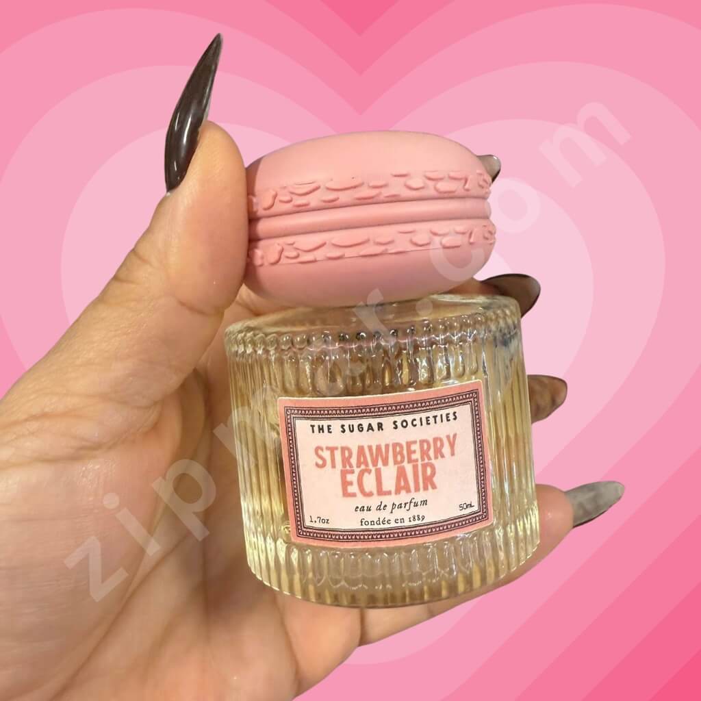 Hand holding a small glass container labeled 'Strawberry Eclair' with a pink lid against a pink heart-shaped background.