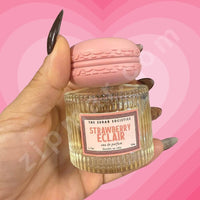 Thumbnail for Hand holding a small glass container labeled 'Strawberry Eclair' with a pink lid against a pink heart-shaped background.